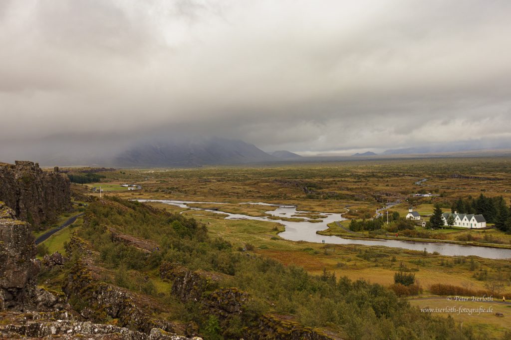 Landschaft mit Fluss, Wiesen und bewölktem Himmel, im Hintergrund Berge.