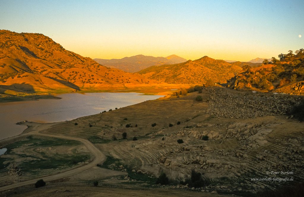 Hügelige Landschaft, mit See, in abendlichem Licht der untergehenden Sonne.