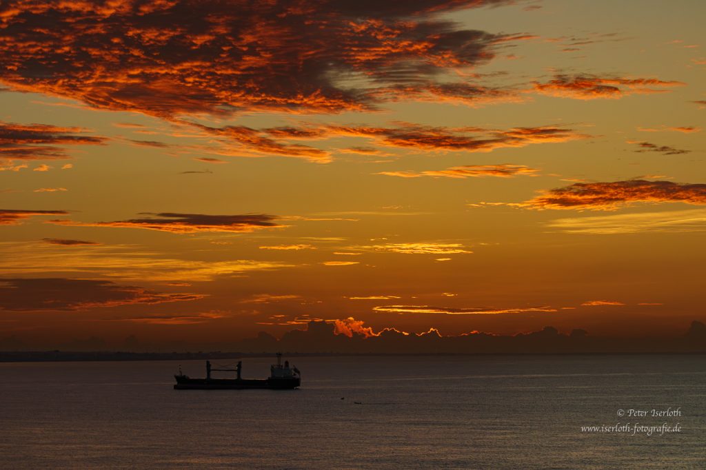 Sonnenaufgang in der Karibik, kurz bevor die Sonne über den Horizont steigt. Rote Wolken und ein Frachtschiff sind zu sehen.