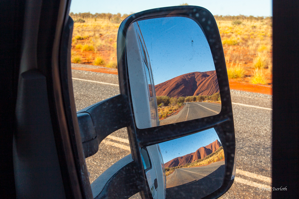 Foto von einem Rückspiegel, der Rücksicht bedeuten soll und den Ayers Rock zeigt.